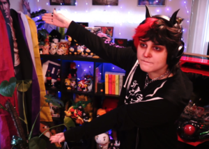 Photo of MomoMisfortune gesturing to a shelf behind them filled with furbies and other collectibles