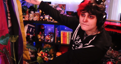 Photo of MomoMisfortune gesturing to a shelf behind them filled with furbies and other collectibles