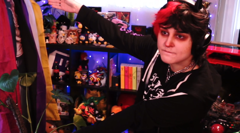 Photo of MomoMisfortune gesturing to a shelf behind them filled with furbies and other collectibles
