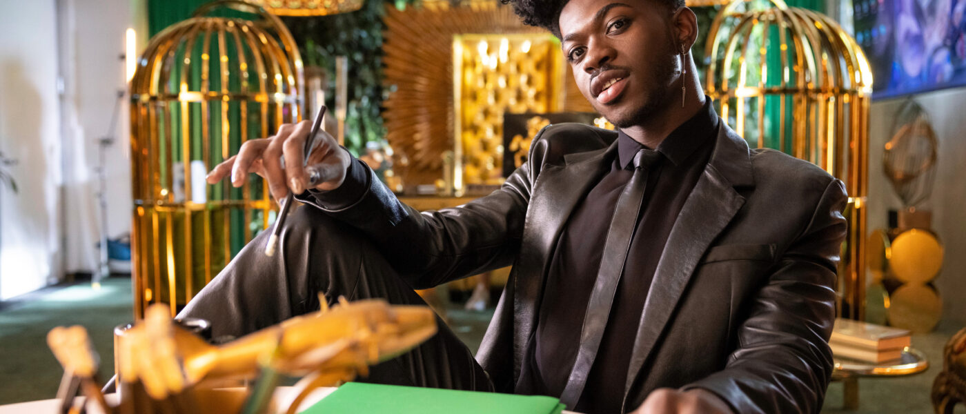 Photo of Lil Nas X sitting at a table in a fully black suit