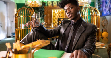 Photo of Lil Nas X sitting at a table in a fully black suit