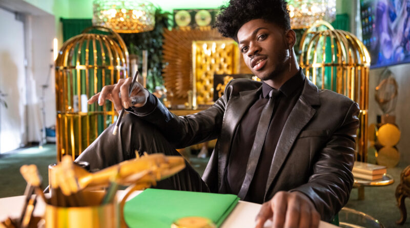 Photo of Lil Nas X sitting at a table in a fully black suit